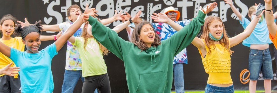 Teenage children with their hands in the air singing outdoors against a black backdrop